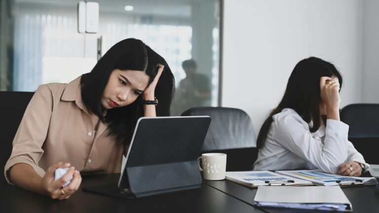 Two frustrated business professionals in office meeting room showing stress and exhaustion, illustrating how leadership identity patterns create dysfunction and bottlenecks in executive teams.